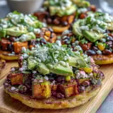 A close-up shows sliced avocado and crumbled feta topping the Black Bean and Sweet Potato Tostadas, garnished with fresh cilantro and a lime wedge.
