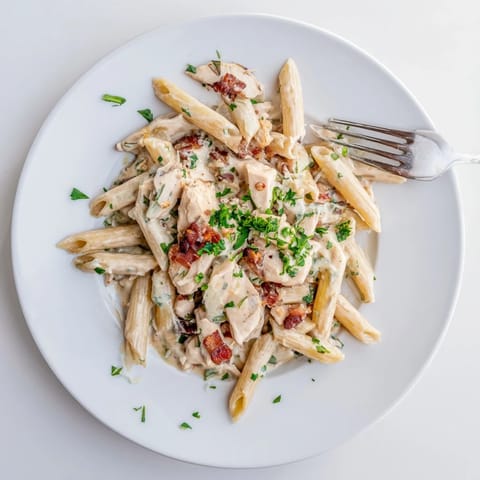 A close-up of Creamy Chicken Bacon Penne, garnished with parsley and served alongside a glass of white wine.  