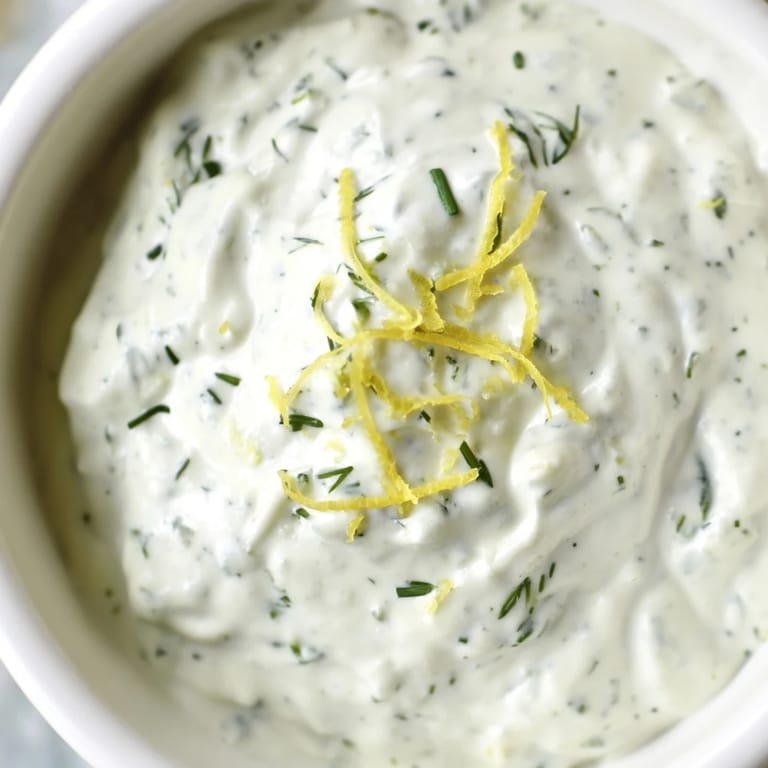 A close-up of Greek Yogurt Herb Dip, thick and tangy, with visible herb flecks, paired with colorful raw veggies for dipping.