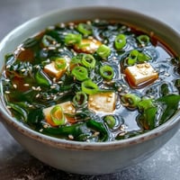 A close-up of warm Wakame Soup with tender seaweed, silky tofu cubes, and fresh green scallions in a savory dashi broth.  